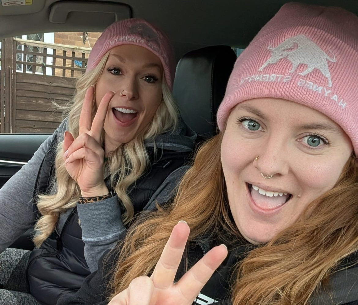 Jess and Lizzie in a car wearing pink beanies, making peace signs.